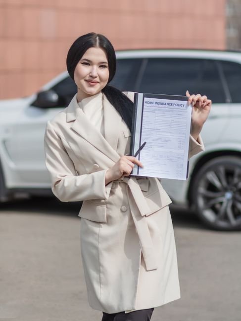 compare.com car insurance - Smiling woman in beige blazer holding a home insurance policy document in outdoor setting.