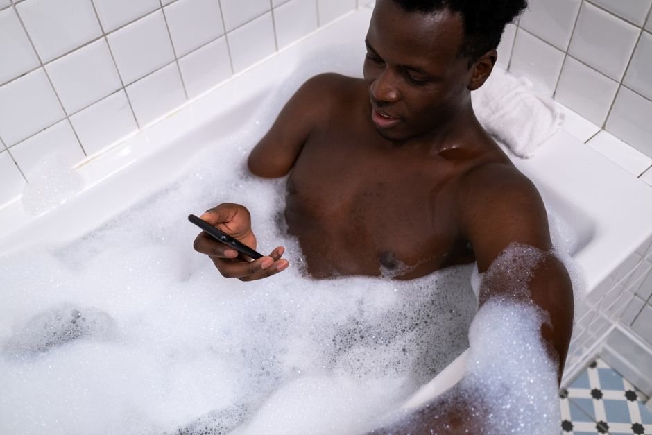 CompareMyQuotes Bathroom CompareMyQuotes Bathroom - An adult man enjoying a bubble bath while using a mobile phone in a white tiled bathroom.